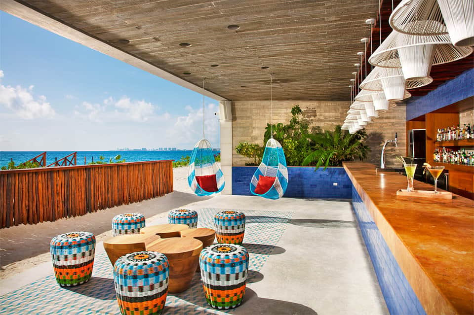 Modern beachfront bar lounge with woven pendant lights, colorful seating, ocean views, and wooden accents