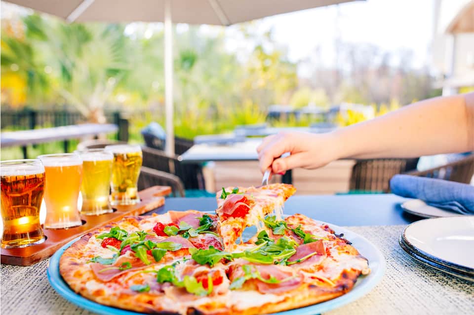 Person eating pizza at outdoor table with beer glasses and tropical garden views at resort