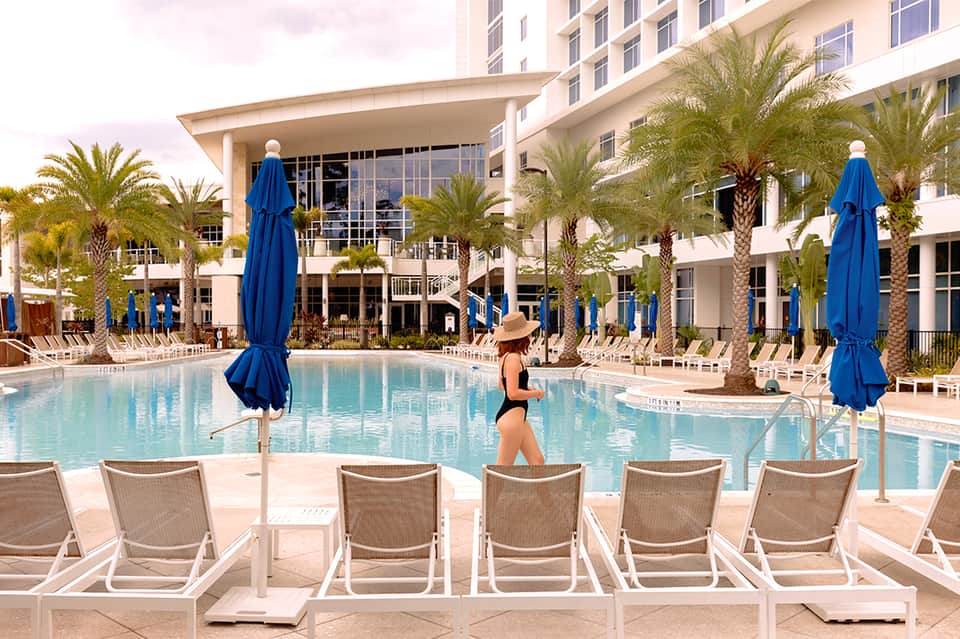 Resort pool with blue umbrellas, lounge chairs, palm trees, and modern white building