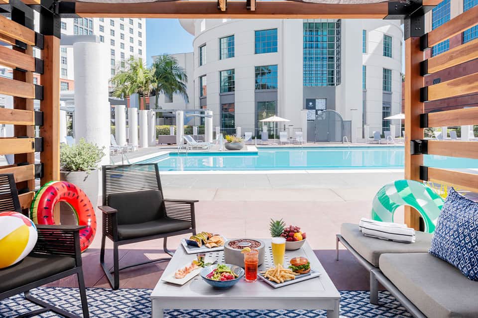 Modern hotel poolside lounge with colorful beach balls, food spread, and turquoise pool beyond