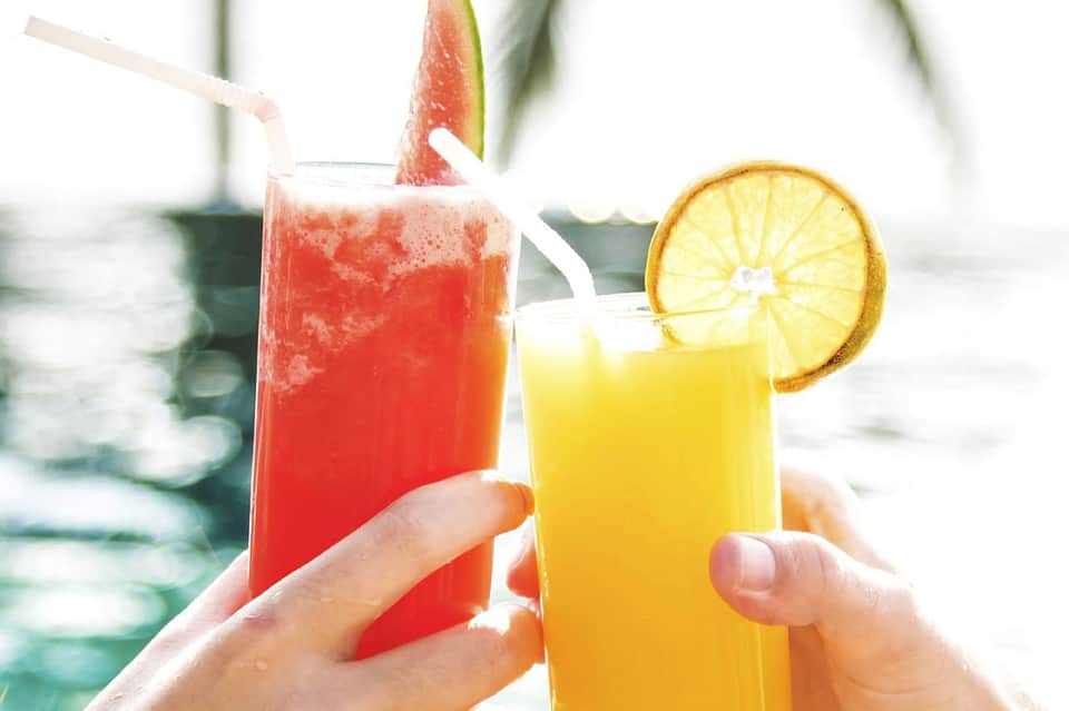 Two tropical drinks held by hands overlooking a swimming pool and beach loungers