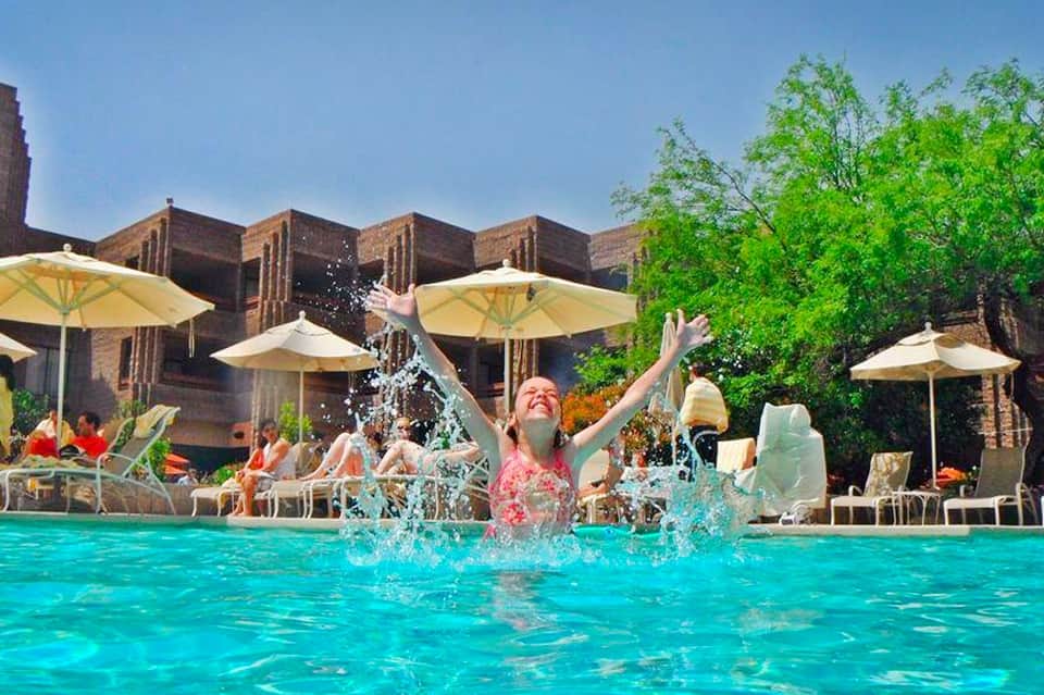 Woman splashing in bright turquoise pool with umbrellas and brick buildings in background