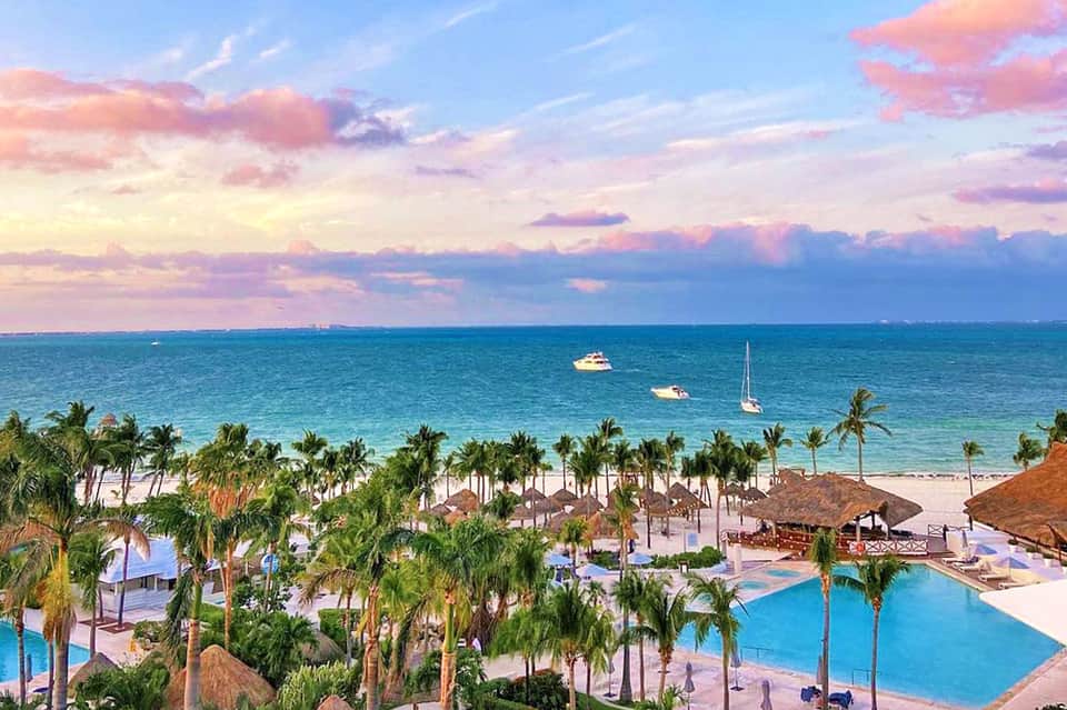 Aerial view of beachfront resort with multiple pools, palm trees, tiki umbrellas, white sand beach, and sailboats on turquoise water
