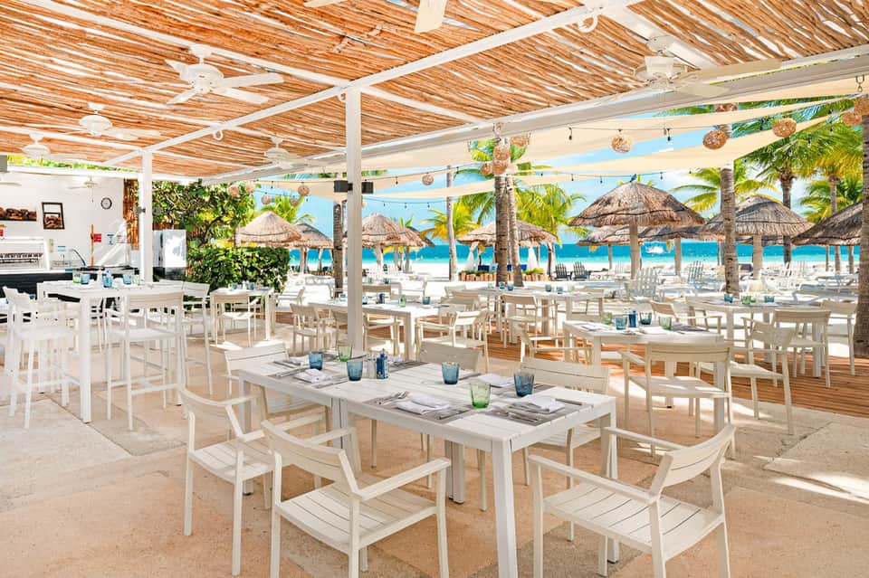 Beachfront restaurant with white furniture, wooden lattice ceiling, thatched umbrellas, and ocean beyond