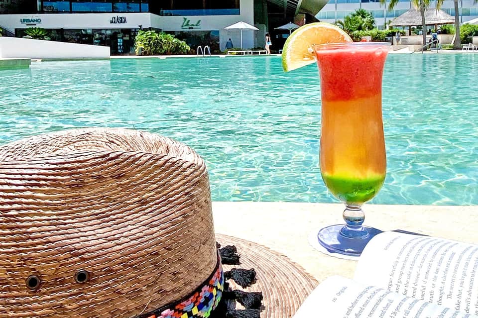Colorful tropical drink with layered colors and orange slice near pool with straw hat