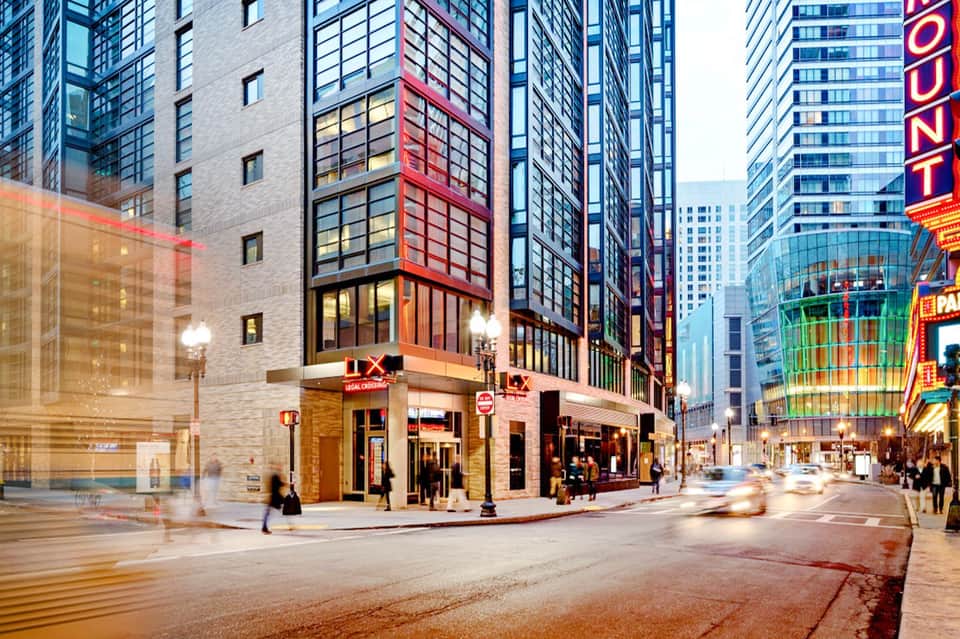 Urban street corner with modern high-rise buildings, ground-level hotel entrance with glass storefront at dusk