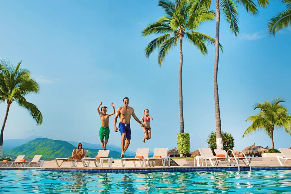 Family jumping into tropical pool with palm trees, mountains, and lounge chairs