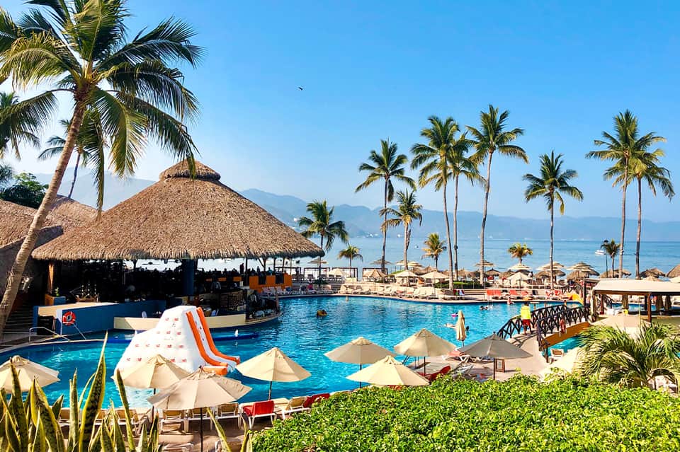 Tropical beachfront resort pool with thatched-roof bar, palm trees, umbrellas, and ocean view in background