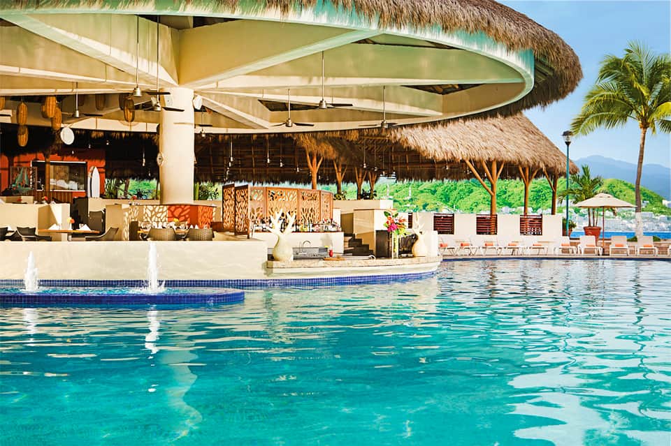Luxury resort pool bar with thatched roof, ocean view, and mountains in background