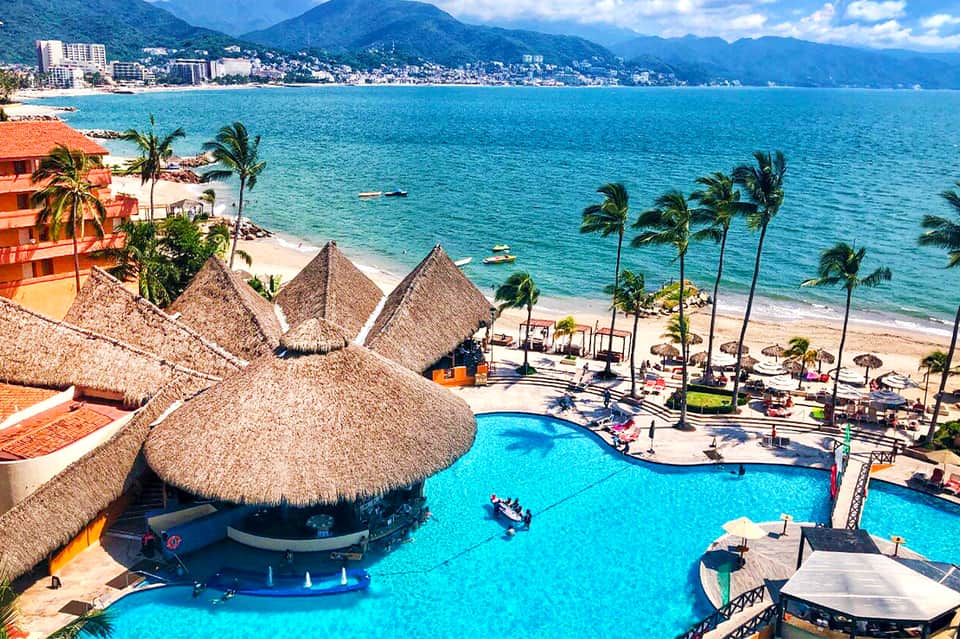 Aerial view of resort pool with thatched palapas overlooking turquoise ocean and beach