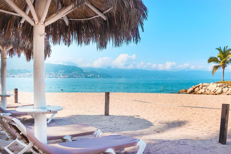 Beach lounge area with thatched umbrella, white lounge chairs, palm trees, and calm ocean waters with distant mountains