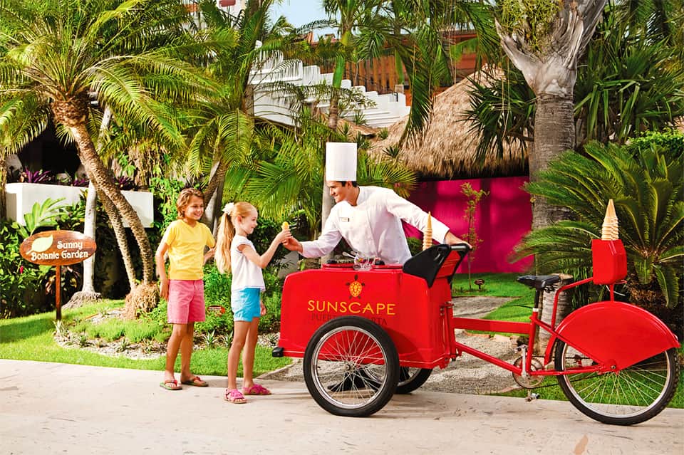 Chef serving ice cream from red Sunscape cart to children in tropical garden