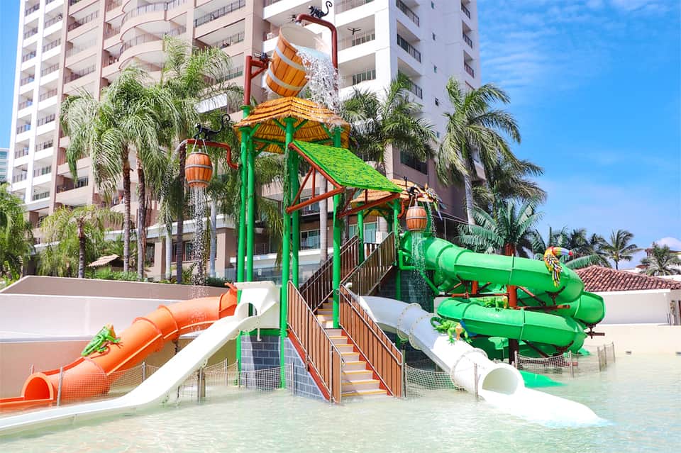 Colorful water park with green and orange slides, splash bucket, and palm trees