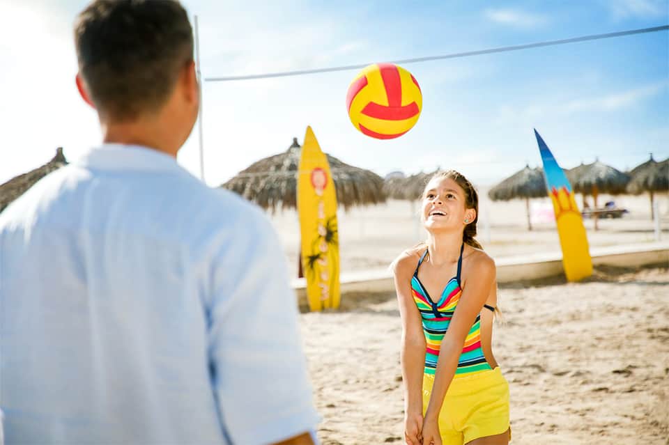 Father and daughter playing with colorful ball on beach with thatched umbrellas in background