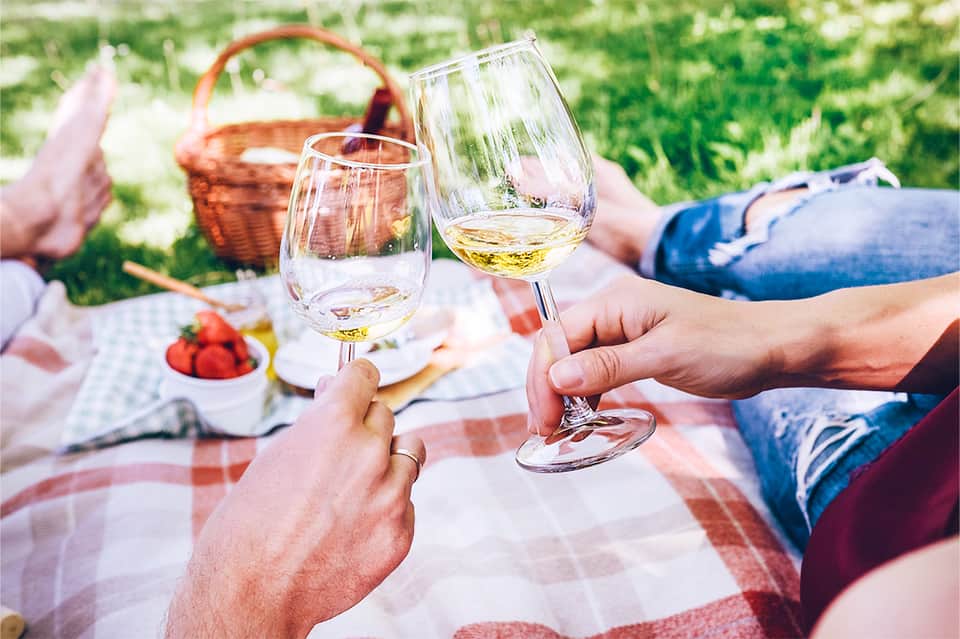 Two wine glasses clinking during an outdoor picnic on a checkered blanket with a basket and strawberries