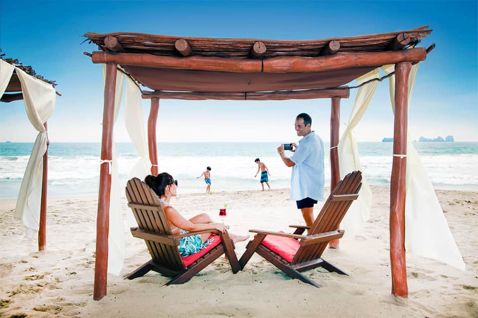 Couple relaxing in wooden beach cabana with white curtains overlooking turquoise ocean