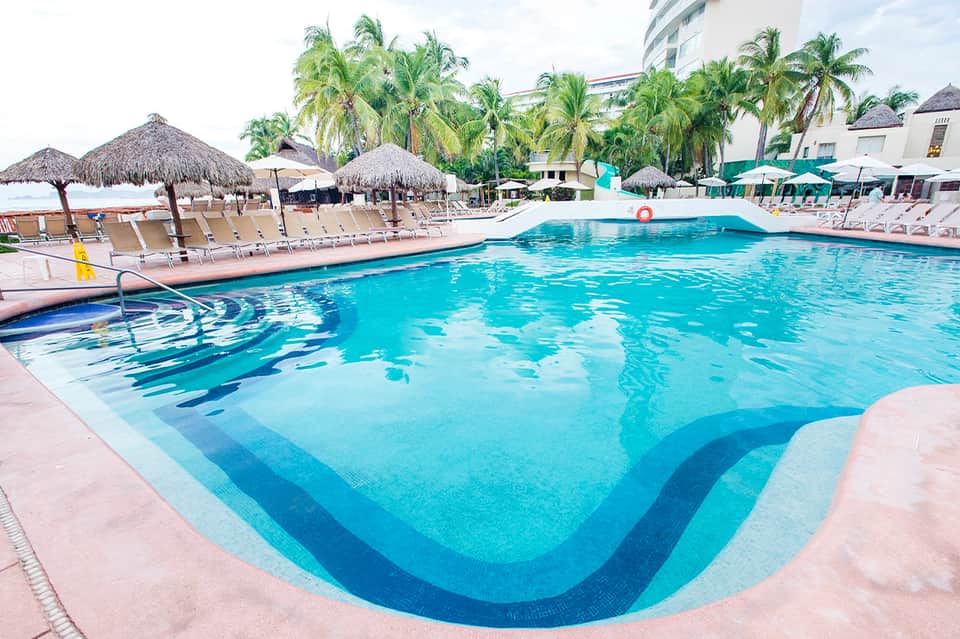 Resort swimming pool with wave design, tiki umbrellas, lounge chairs, and palm trees lining the deck