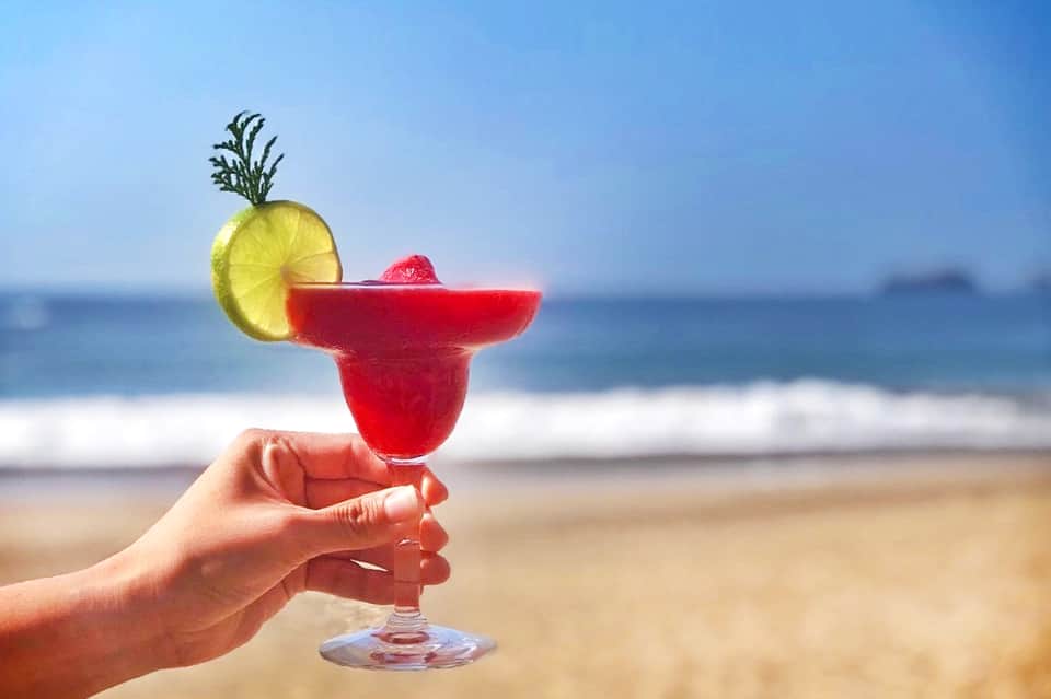 Hand holding red margarita with lime slice and pineapple against sandy beach and ocean
