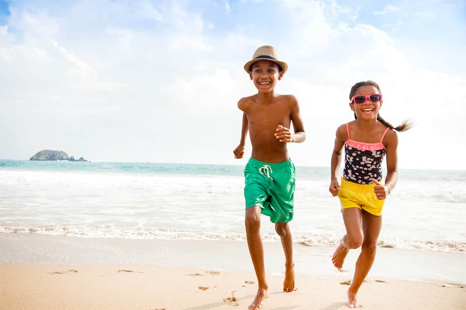 Two children running on sandy beach in bright sunlight with clear turquoise ocean water behind them