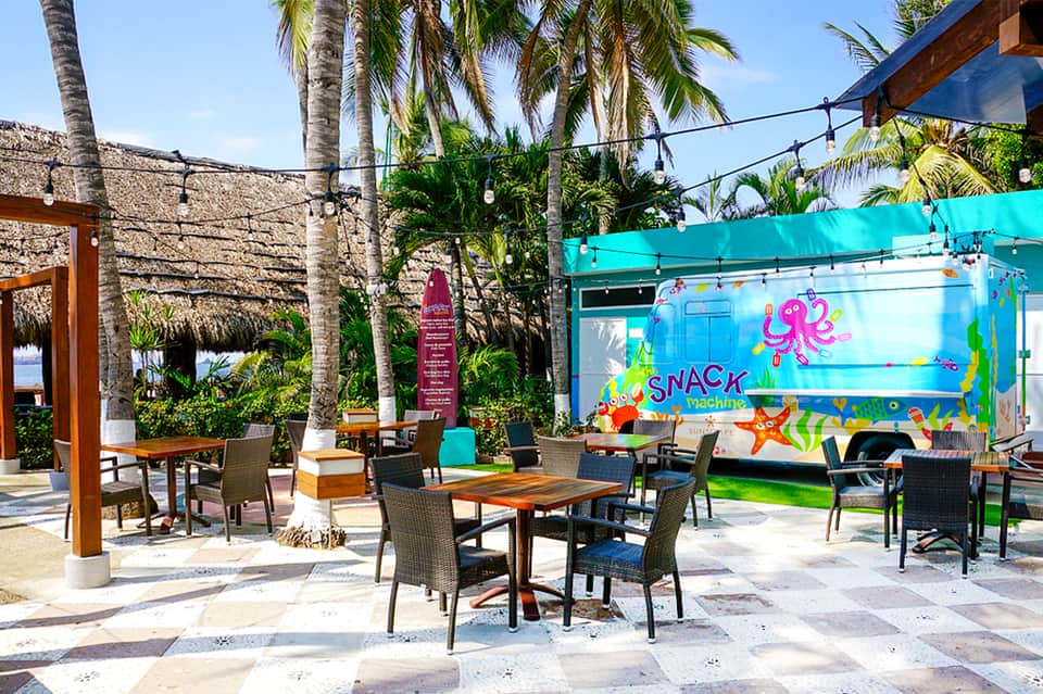Outdoor tropical resort area with colorful snack machine, palm trees, and casual seating