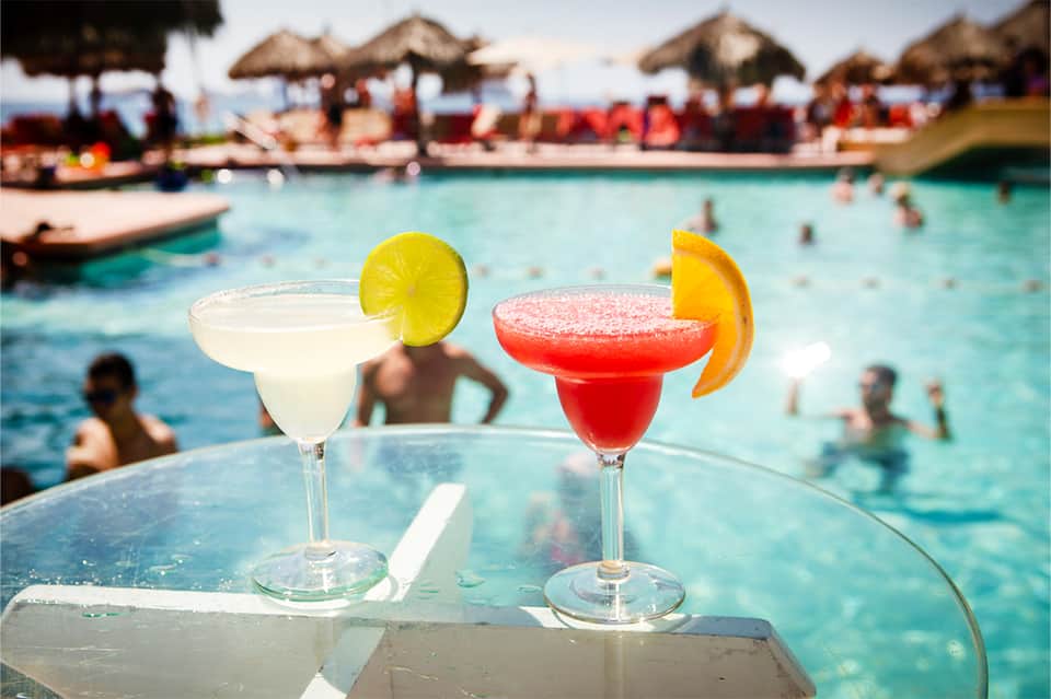 Two tropical cocktails with citrus garnish overlooking beach pool and cabanas