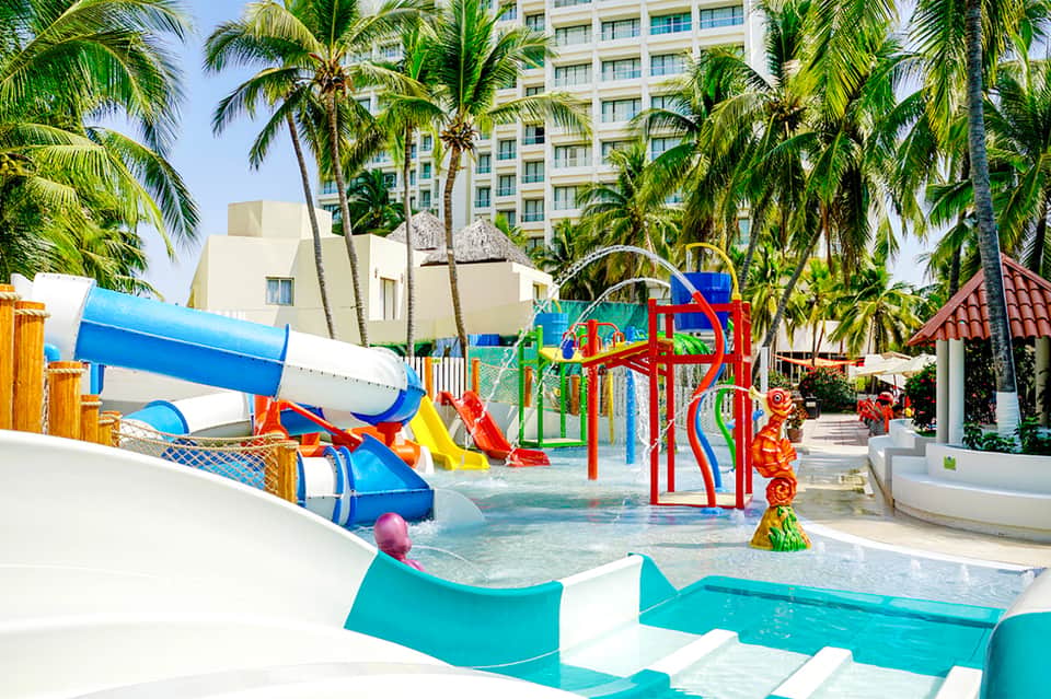Colorful water park with slides, shallow pools, and palm trees near resort buildings