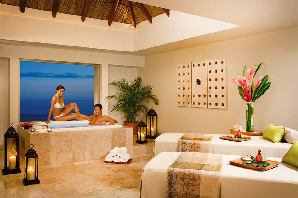 Luxury spa room with jacuzzi tub, massage beds, plants, and ocean view through large window