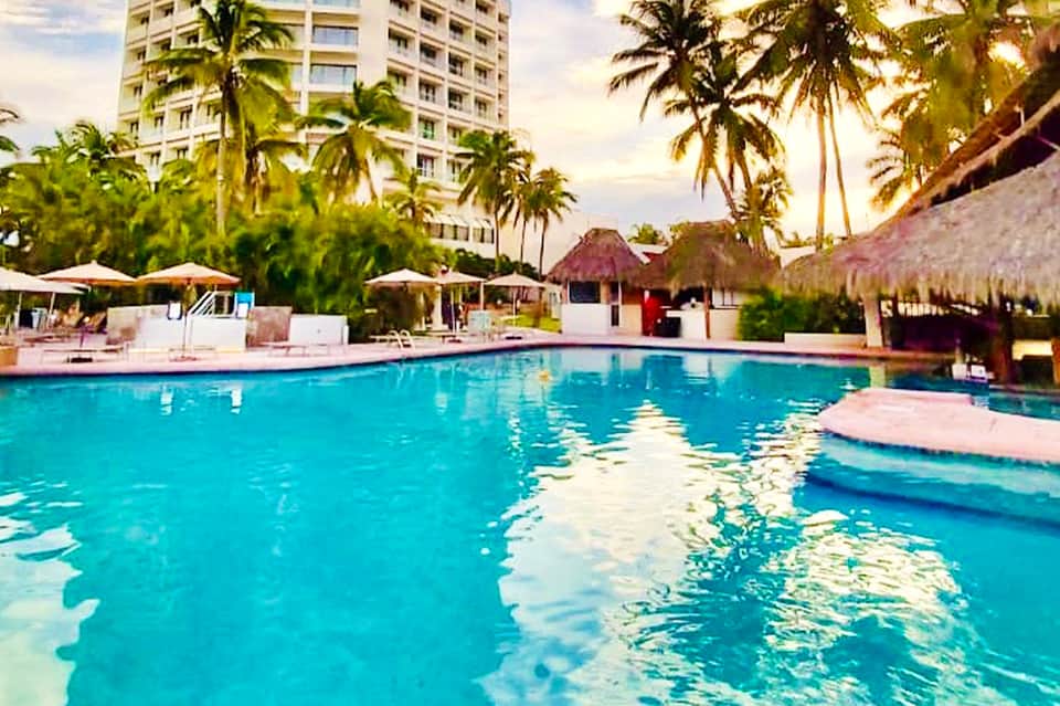 Resort pool surrounded by palm trees, white umbrellas, lounge chairs, and white high-rise hotel building