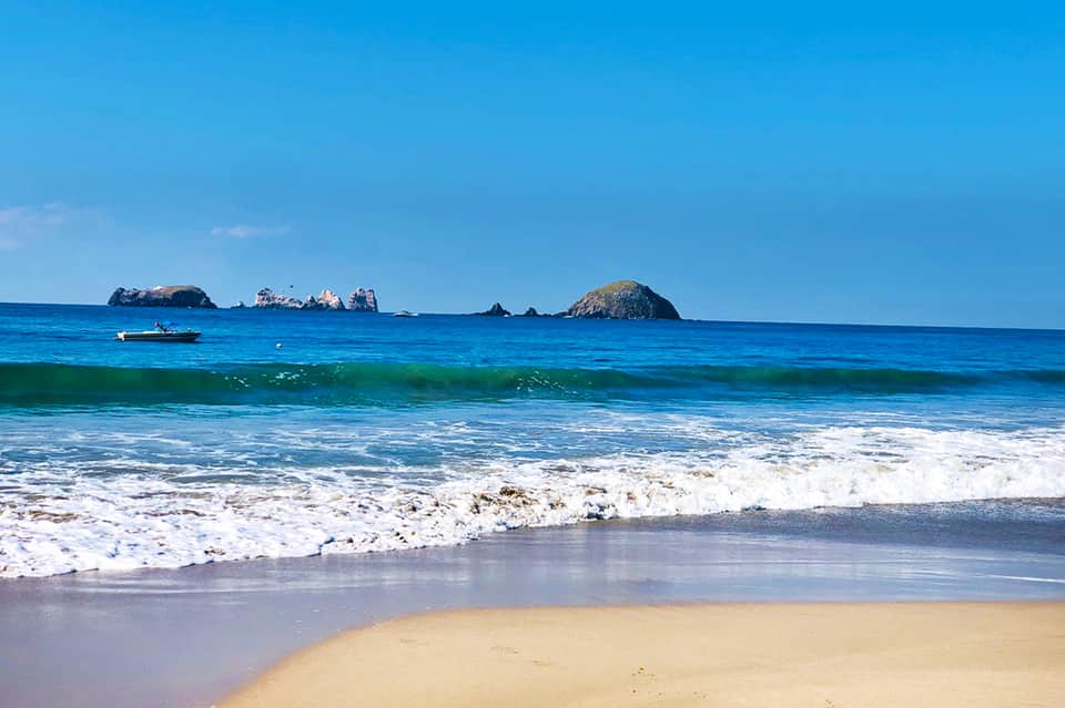 Scenic beach view with rocky islands, blue ocean, white sandy shore, and crashing waves