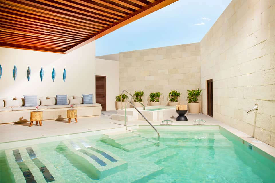 Modern indoor spa pool with turquoise water, lounging area, and stone walls