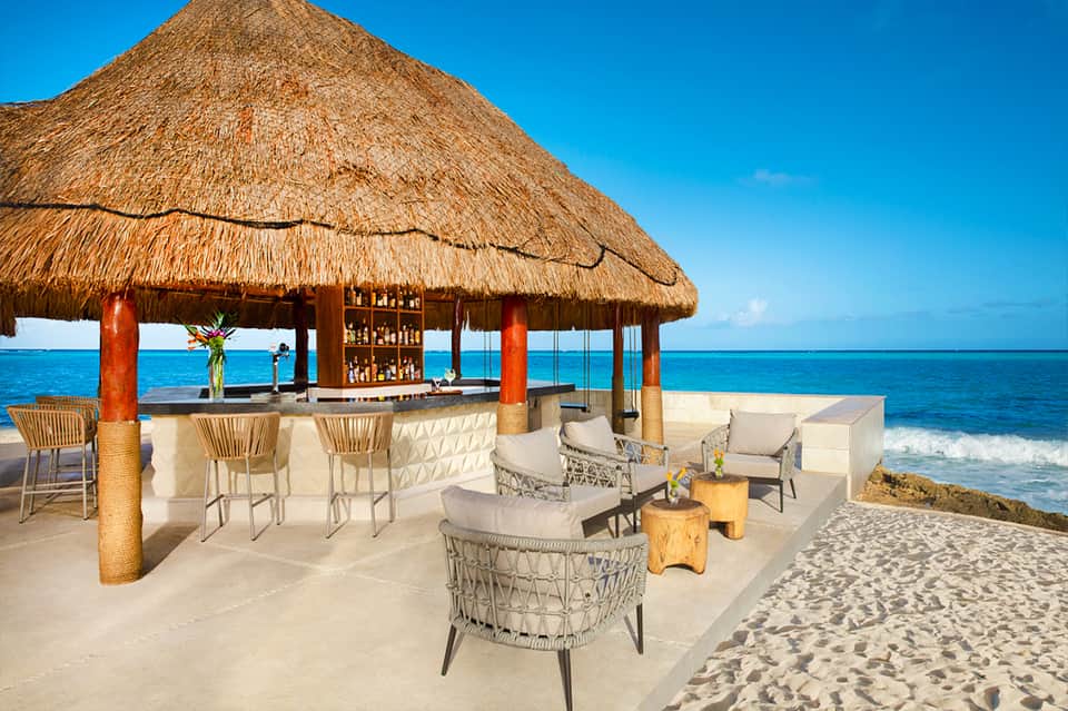 Beachfront tiki bar with thatched roof, lounge seating, and turquoise ocean view