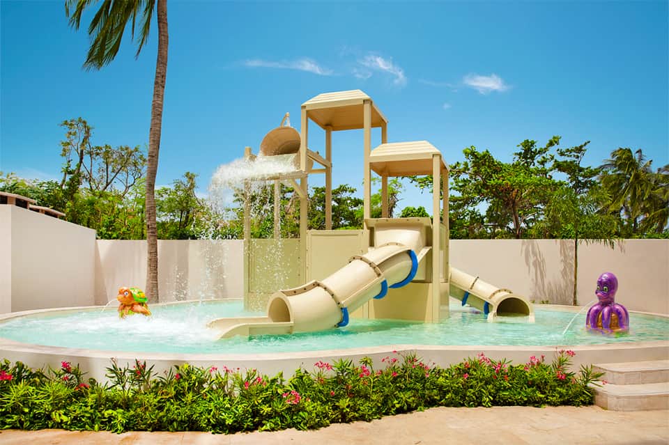 Kids' water play area with beige slide tower, water features, tropical landscaping, and clear shallow pool