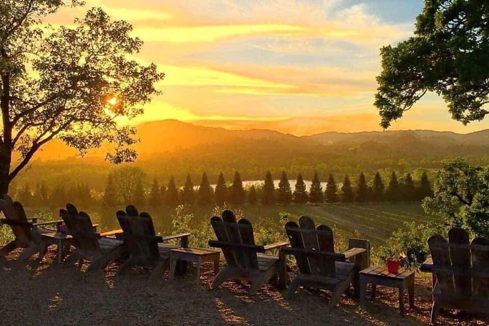 Adirondack chairs facing sunset over vineyard with mountains and water