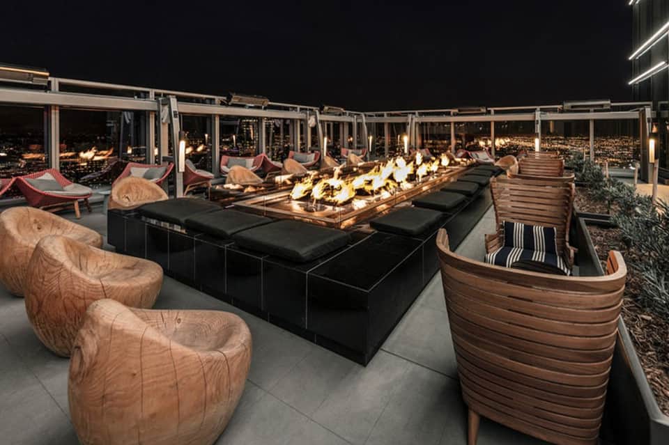 Upscale rooftop lounge at night with fire pit, wooden seating, and city lights view