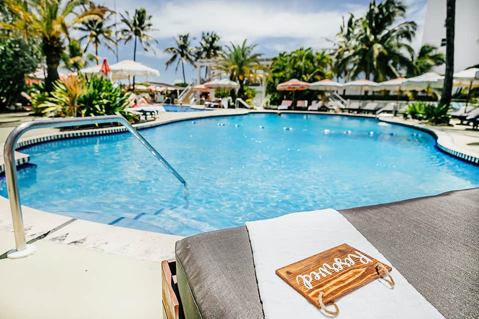 Resort pool area with lounge chairs, palm trees, white umbrellas, and mountain views in background