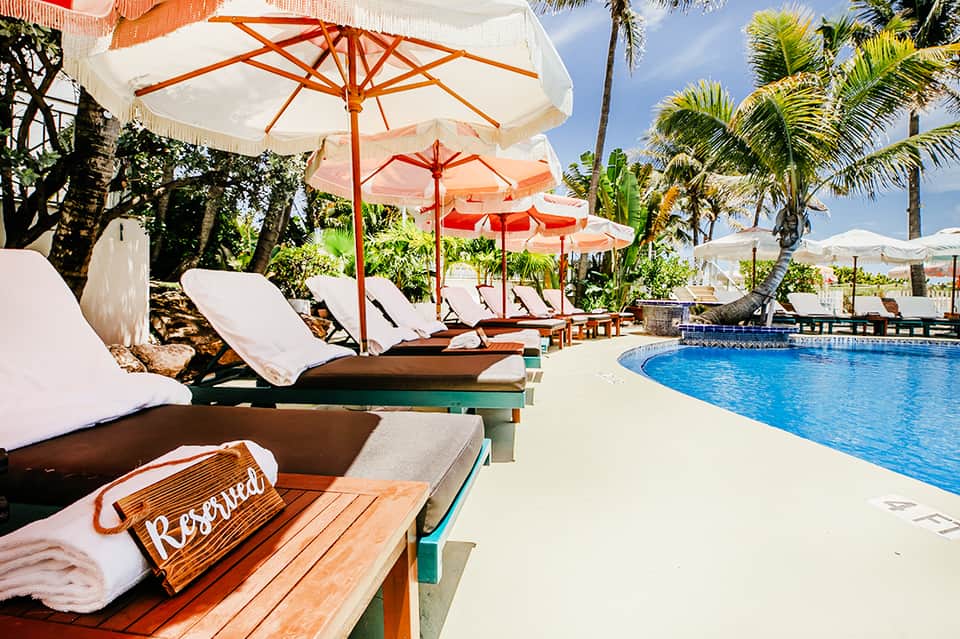 Poolside lounge area with white cushioned chairs under red and white umbrellas among palm trees