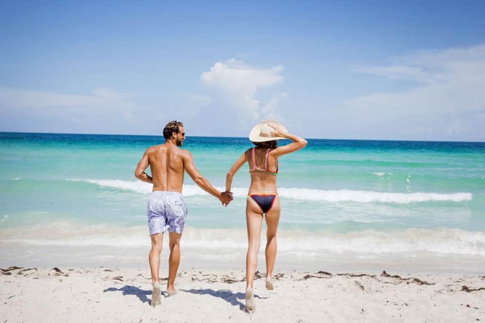Couple holding hands running into turquoise ocean waves on sandy beach