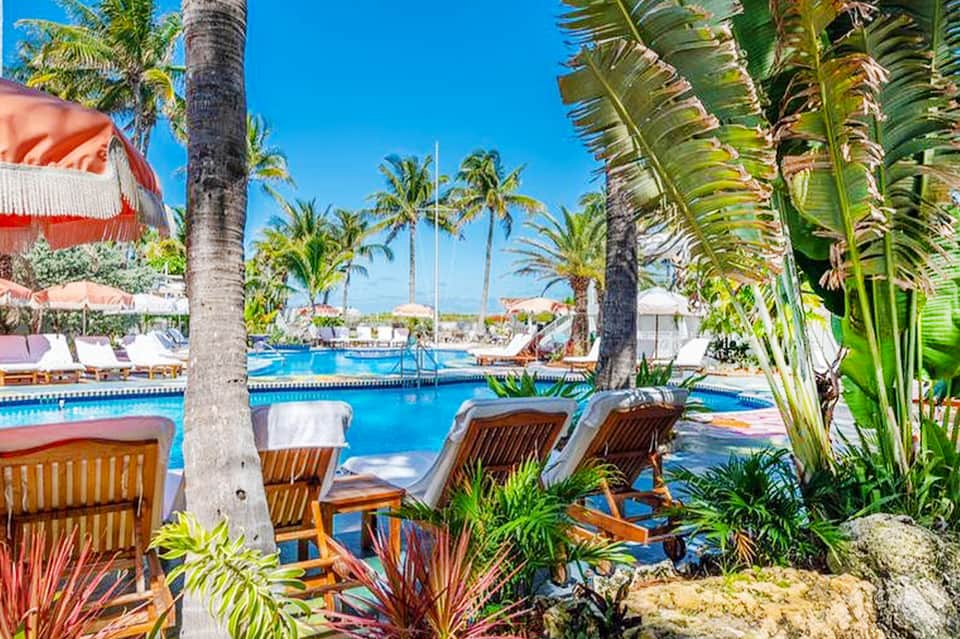 Resort pool surrounded by palm trees, wooden lounge chairs, red and white umbrellas, and clear sky