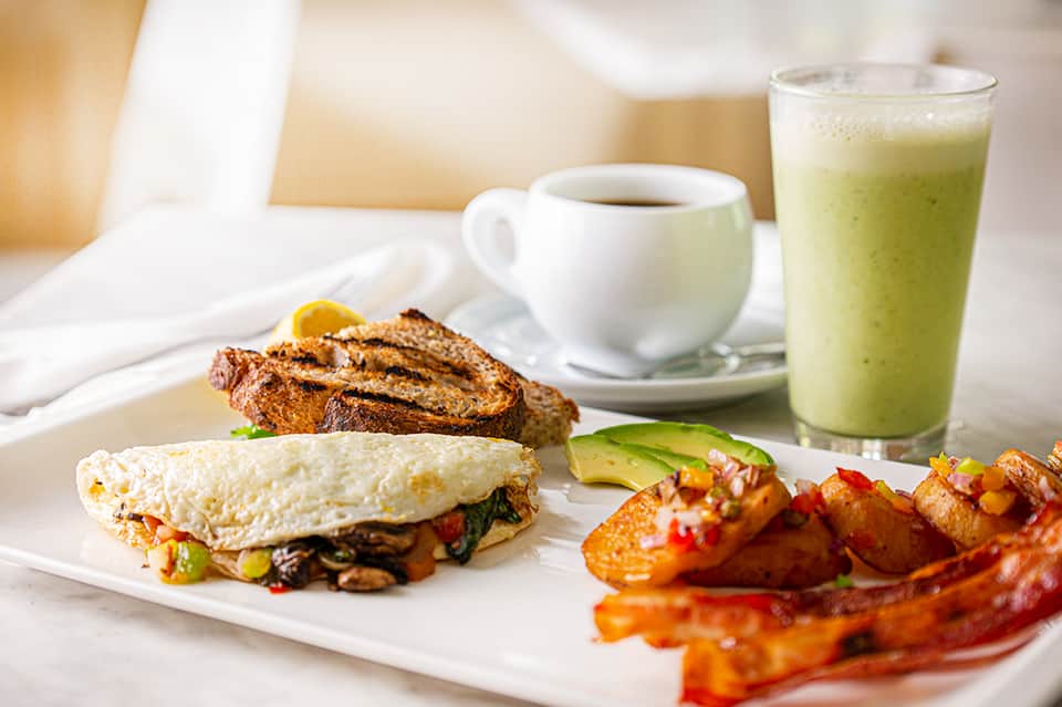 Breakfast plate with omelet, bacon, toast, coffee, and green juice on white table