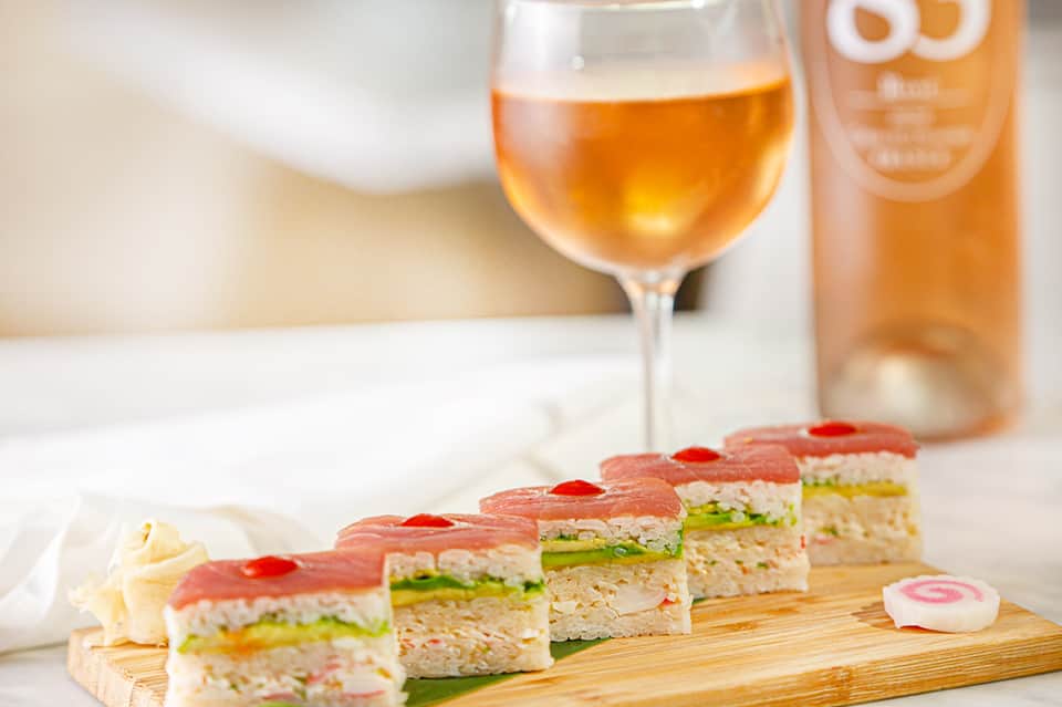 Gourmet tea sandwiches on wooden board with rosé wine and bottle on white surface