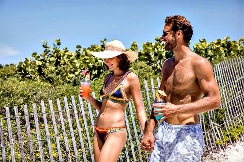 Couple in swimwear enjoying tropical drinks on beach with vineyard in background