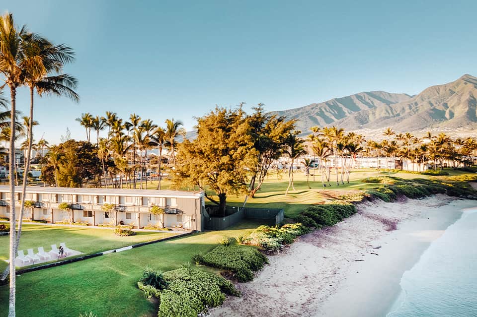 Beachfront resort grounds with green lawn, palm trees, sandy beach, and mountains in the distance