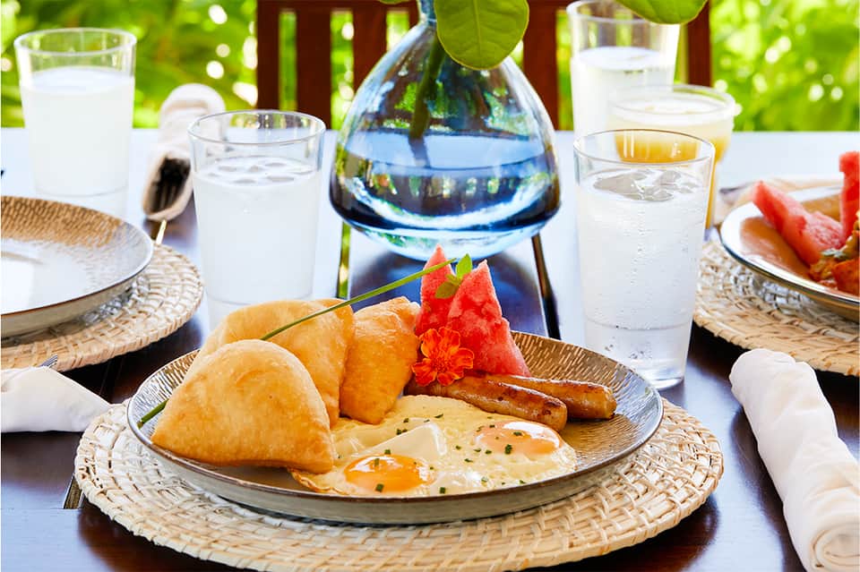 Outdoor dining table set with tropical breakfast including pastries, fruit, eggs, beverages, and ocean view backdrop