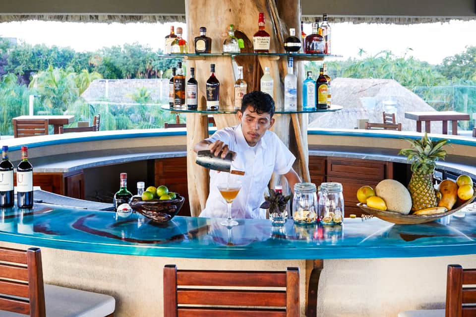 Bartender pouring cocktail at outdoor tiki bar overlooking tropical landscape and palm trees