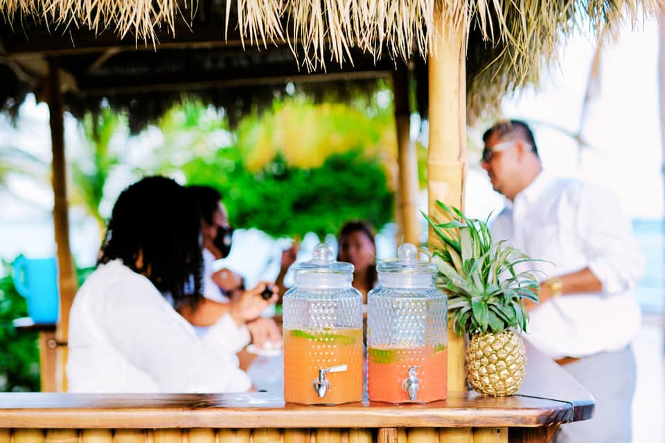Beachside bar with juice dispensers and pineapple, guests dining under thatched roof structure