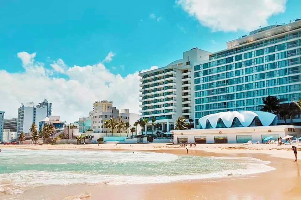 Miami beachfront with modern high-rise hotel buildings, turquoise water, sandy beach, and blue sky.