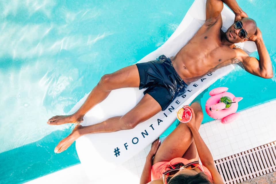 Man relaxing on float raft in turquoise pool with tropical drinks and pink floaties