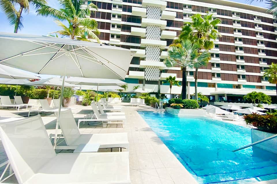 Resort pool with white umbrellas, loungers, high-rise hotel building, and palm trees under clear blue sky