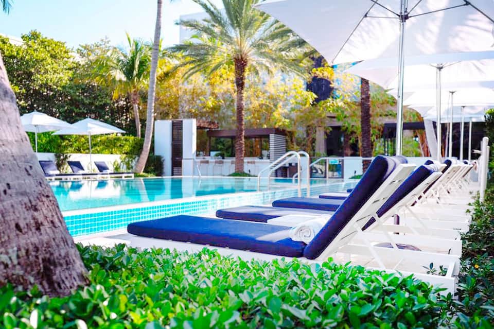 Luxurious resort pool with blue loungers, white umbrellas, palm trees, and modern white buildings