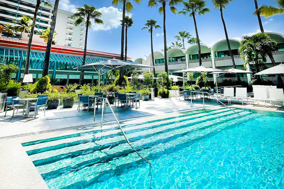 Outdoor lap pool with lane dividers, palm trees, modern building, and shaded seating areas